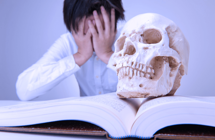 Person looking distraught with head in hands, a fake skull or a human head sits on a book in the forefront.