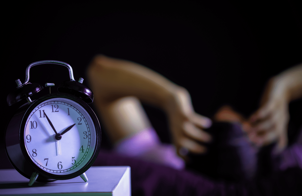 An alarm clock in the forefront, someone holding their hands over their face in the background.